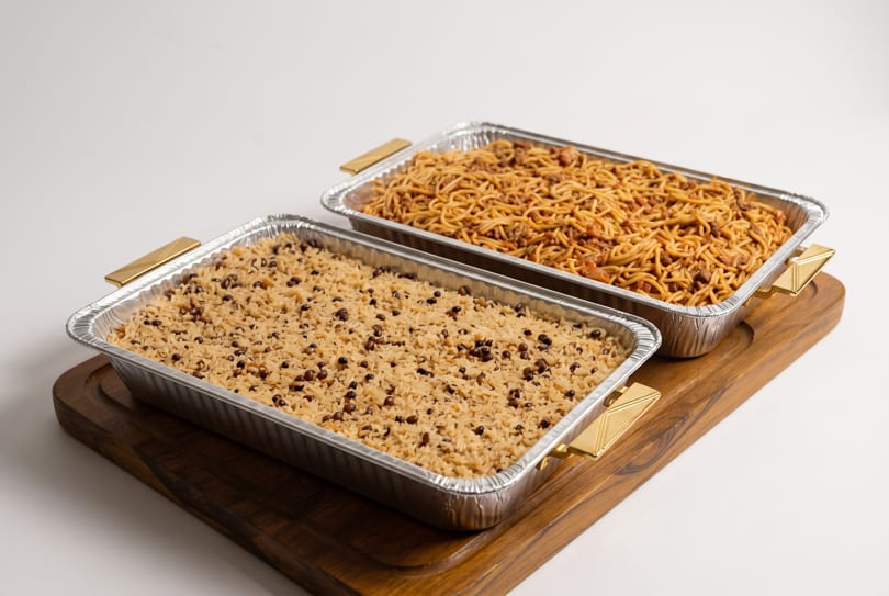 Two catering trays, one with rice and black beans, the other with spaghetti, on a wooden board.