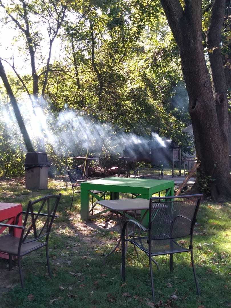Outdoor dining area with green and red tables, chairs, and smoke from a grill under sun-dappled trees.