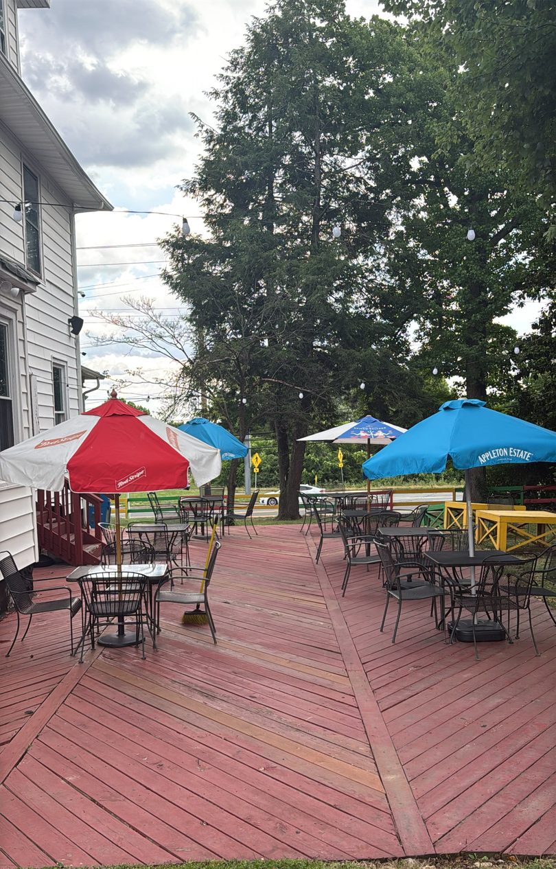 Outdoor patio with red wooden deck, black metal tables and chairs, and colorful umbrellas under trees.