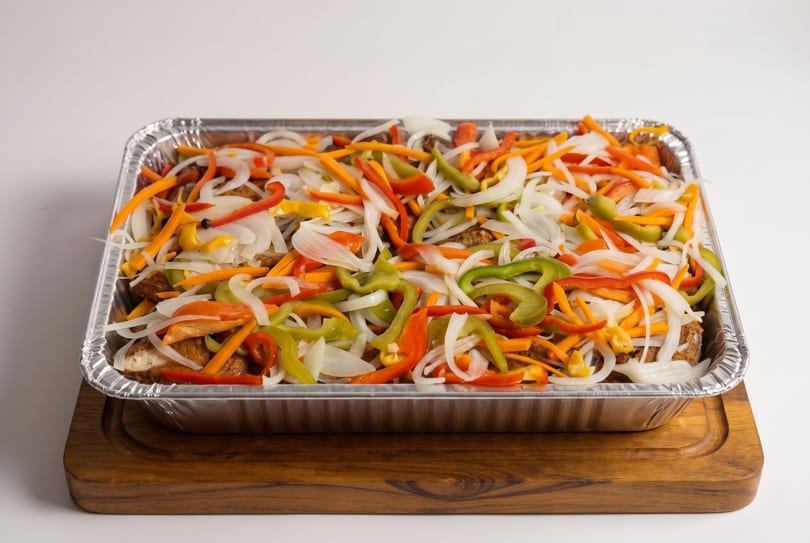 A foil pan filled with colorful sliced bell peppers and onions over meat, on a wooden board.