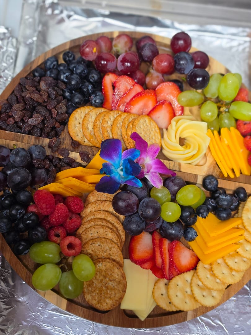 A wooden charcuterie board filled with crackers, various cheeses, fresh berries, grapes, and mango slices.