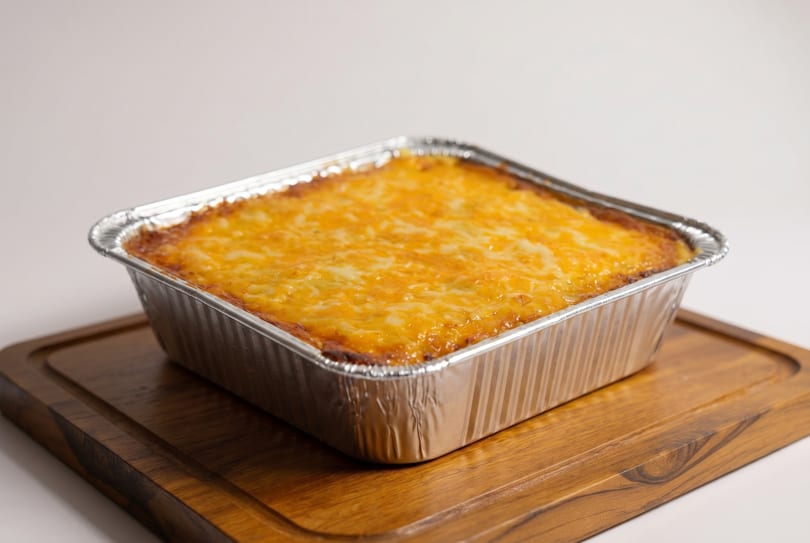 A rectangular aluminum foil pan filled with a cheesy baked dish, resting on a wooden board.