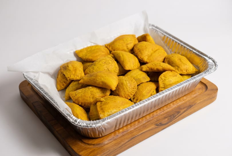 A foil tray filled with numerous golden-yellow Jamaican beef patties, lined with white parchment paper.