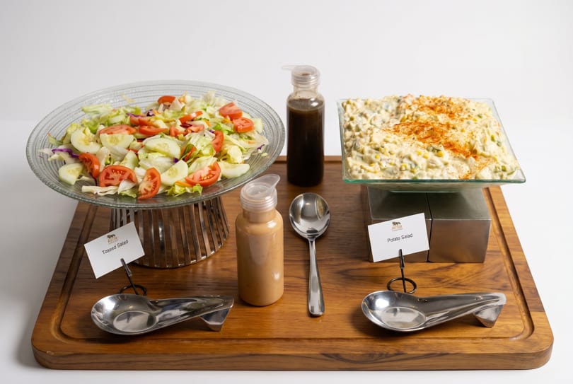 Tossed salad and potato salad with dressings and serving spoons on a wooden board.