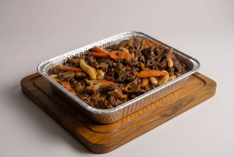 Oxtail stew with carrots and beans in a foil pan on a wooden cutting board.