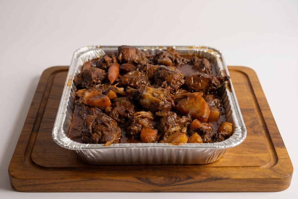 Hearty beef stew with potatoes and carrots in a foil pan on a wooden cutting board.